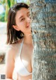 A woman in a white bikini leaning against a palm tree.