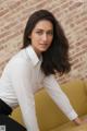 A woman in a white shirt and black pants leaning against a brick wall.