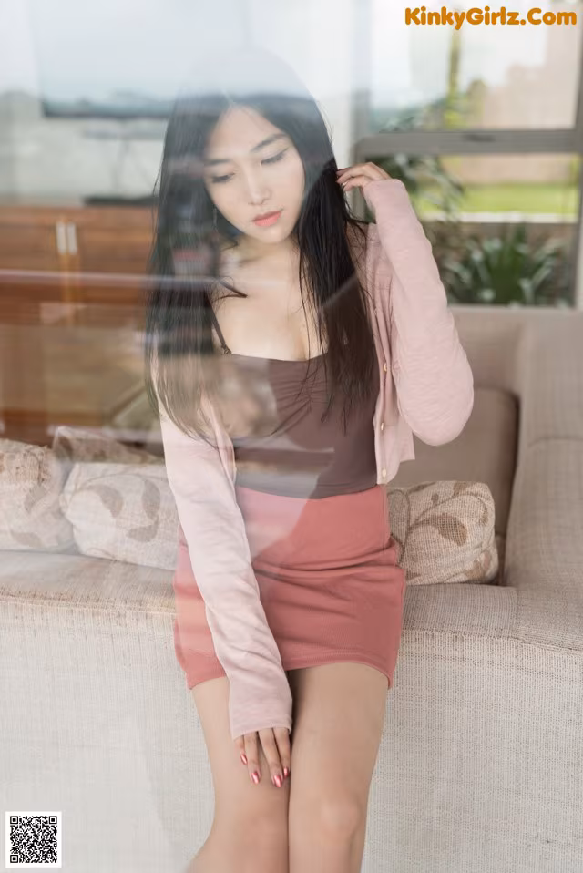 A woman in a pink skirt and a pink cardigan posing on a couch.