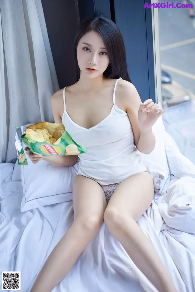 A woman sitting on a bed holding a bag of chips.