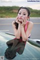 A woman leaning on the hood of a car with her hand on her chin.